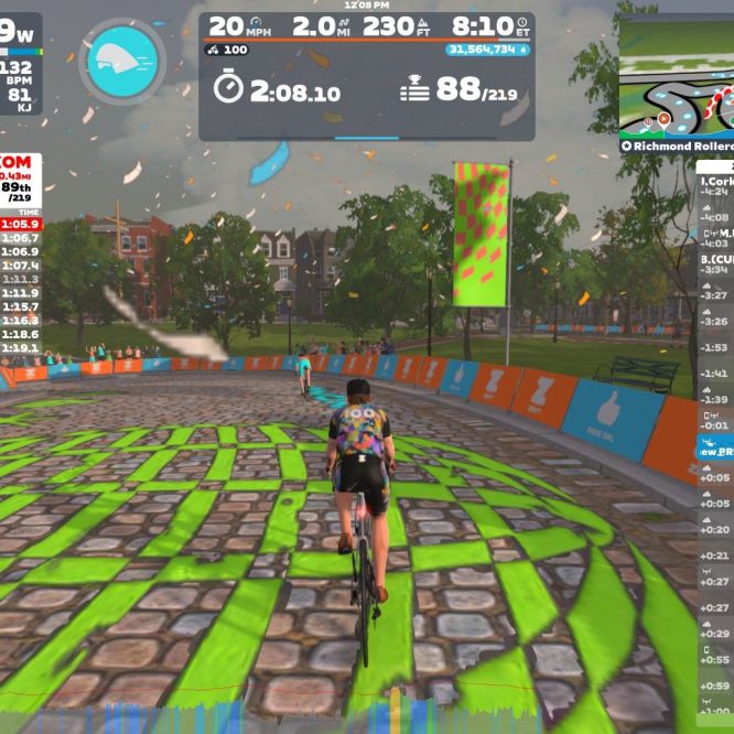A cyclist navigates a colorful virtual course in Zwift, showing speed and power metrics on the screen. In the background, people are cheering, and the course features green and striped patterns along the cobblestone path. Various performance statistics and rankings are displayed on the side of the screen.