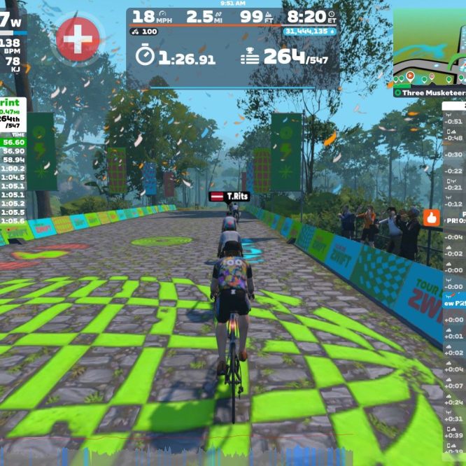 A screenshot from a Zwift cycling game shows a cyclist racing on a colorful, virtual road marked with green patterns. The screen displays cycling stats such as speed (18 mph), power (167 W), and heart rate (138 bpm). In the background, trees line the path, and virtual spectators can be seen cheering. Live results and rankings for the Dos d’Ane Sprint event are visible on the left side of the screen.
