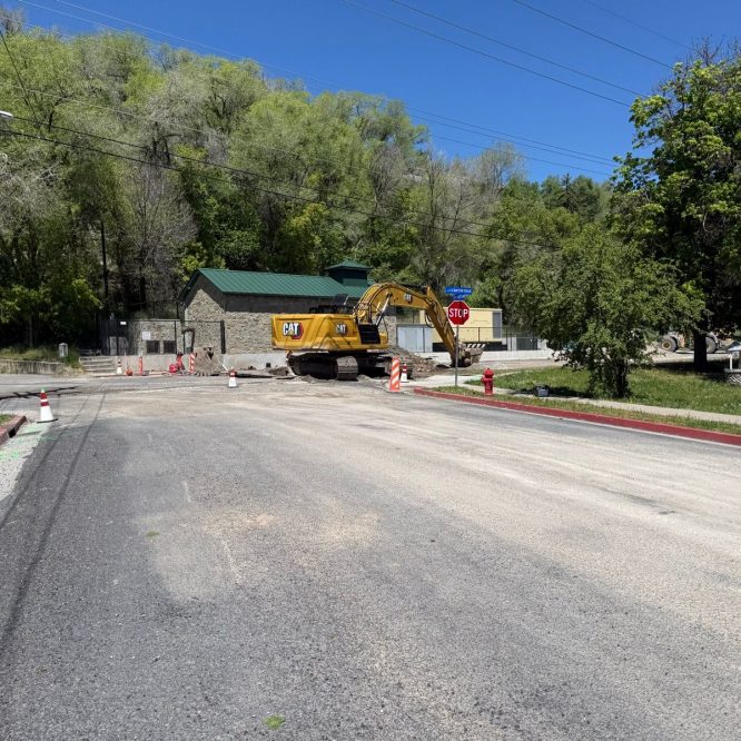 A construction site in Paradise, Utah, featuring a large yellow excavator parked on a newly laid asphalt road. There are traffic cones and a "STOP" sign visible. In the background, a stone building and trees are seen, under a clear blue sky.
