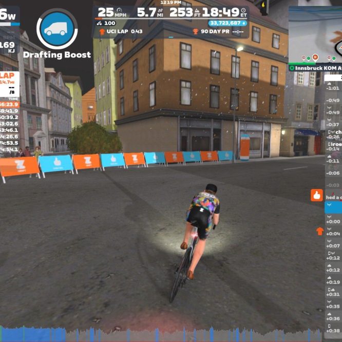 A cyclist is riding on a virtual course in Zwift, wearing a colorful jersey. The screen displays various performance metrics, including power output, speed, and lap times. In the background, there are buildings and barriers marked with the Zwift logo, indicating a competitive cycling environment.