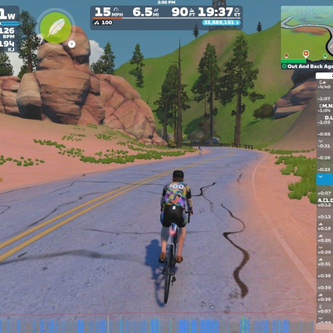 A cyclist wearing a colorful jersey rides on a virtual road in Zwift, surrounded by green trees and rock formations. The screen displays performance metrics, including wattage, speed, heart rate, and distance traveled.