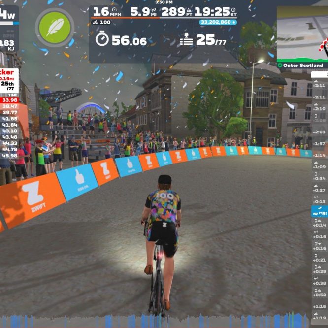 A cyclist rides on a virtual track in Zwift, surrounded by spectators cheering on the sidelines. The cyclist is referencing live race results displayed on the screen, showing rankings and times for participants. The background features colorful banners and elements typical of an outdoor racing environment, while confetti falls from above, adding to the festive atmosphere.
