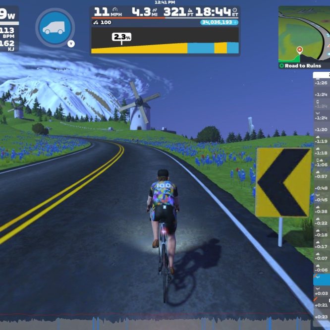 A cyclist rides on a road with a slight incline, surrounded by green hills, blue flowers, and windmills under a clear sky. The screen displays real-time cycling metrics, including power, speed, and distance, along with a leaderboard of nearby riders in the Zwift virtual cycling environment.