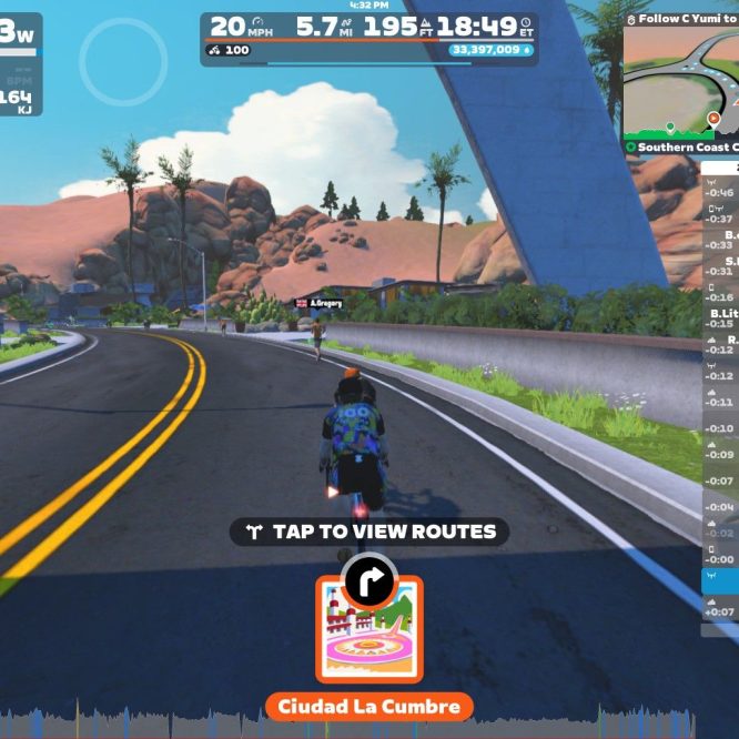 A virtual cycling scene from Zwift, showing a bike rider on a smooth, winding road. The landscape features rocky hills and greenery under a clear blue sky. On-screen stats display power, speed, and distance, along with nearby cyclists’ names and times. A route selection button is visible at the bottom. The location is labeled "Ciudad La Cumbre."