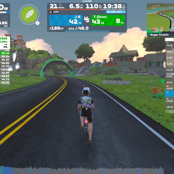A cyclist participates in a virtual race on Zwift, riding on a winding road in a colorful, cartoon-like landscape. The display shows various statistics, including power output, speed, and race standings, with surrounding scenery featuring hills, trees, and buildings.