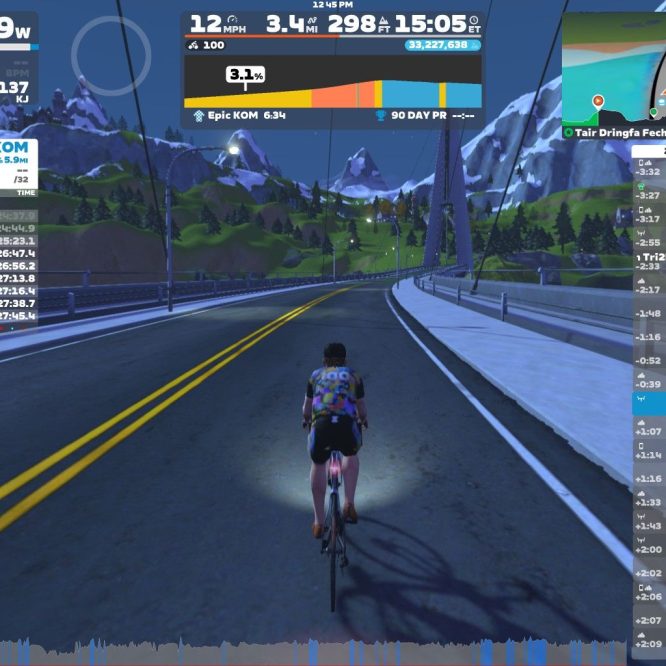A cyclist is riding on a virtual road with mountains in the background, part of the Zwift platform. The screen displays various performance metrics, including power output, speed, distance, and a leaderboard with rankings and times. There is also a map indicating the cyclist's location within the virtual environment.