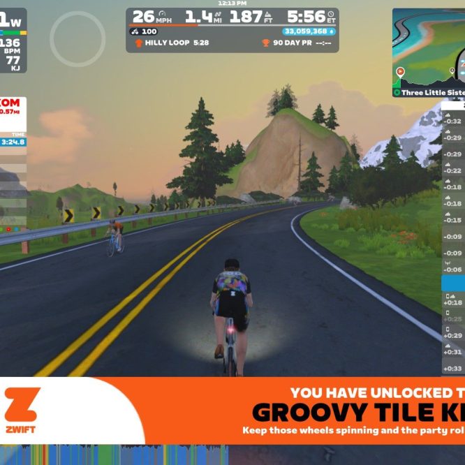 A screenshot from Zwift, an online cycling platform. The scene shows two virtual cyclists riding on a winding road with mountains and trees in the background. Important statistics such as power output, speed, and heart rate are displayed on the screen, along with leaderboard information and a notification about unlocking a new item.