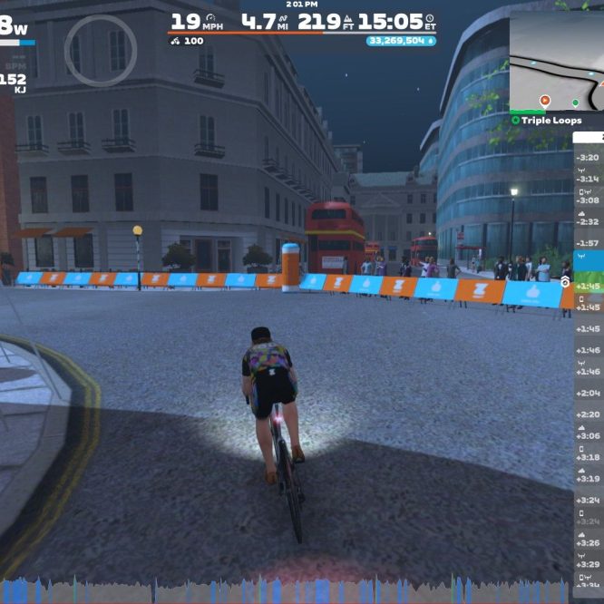 A cyclist is riding on a virtual course in Zwift during a nighttime setting. The cyclist is shown from behind, with a focus on stats like power output and speed displayed on the screen. Surrounding the cyclist are virtual buildings, a road lined with barriers, and a map indicating the course layout. Several other participants are visible nearby, and a double-decker bus can be seen in the background.