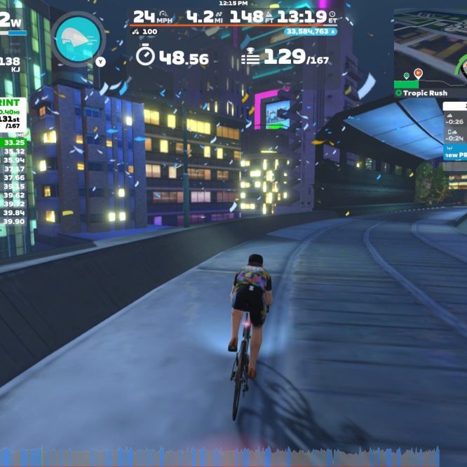 A cyclist rides on a virtual track in Zwift, surrounded by colorful buildings in a nighttime cityscape. The screen displays various statistics, including the cyclist's speed, power output, and a leaderboard with live results of participants in a railway sprint event.