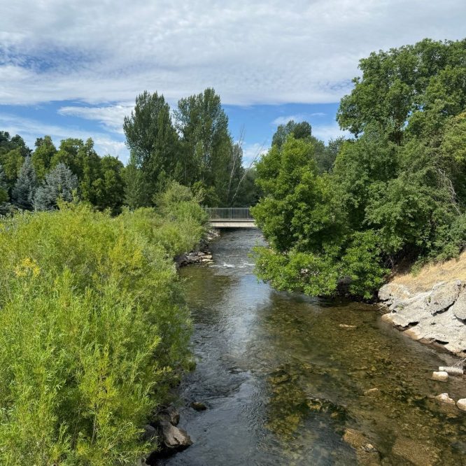 A clear river flows through lush greenery, with trees lining the banks and a bridge visible in the background under a partly cloudy sky. The scene captures a peaceful, natural setting in Paradise, Utah.