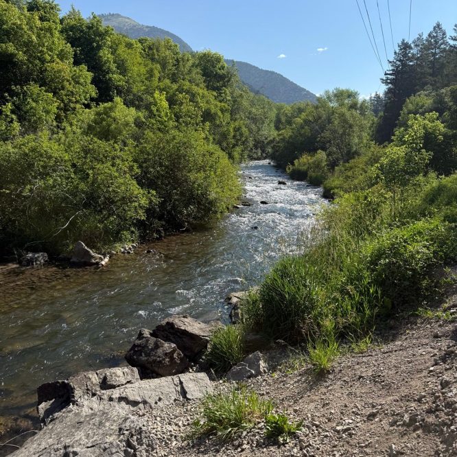 A river flows through a green landscape in Providence, Utah. The scene features lush trees along the riverbanks, rocky areas, and distant mountains under a clear blue sky. Power lines are visible in the background.