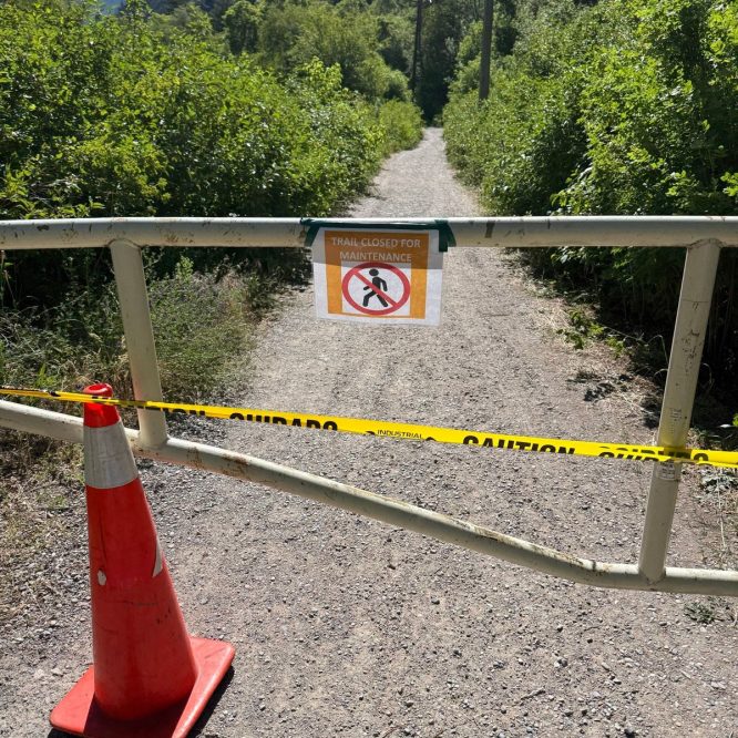 A gravel trail is closed off by a white gate with a sign that reads "Trail Closed for Maintenance." There is a traffic cone on the ground and yellow caution tape around the gate. Lush greenery is visible on either side of the path, with mountains in the background.