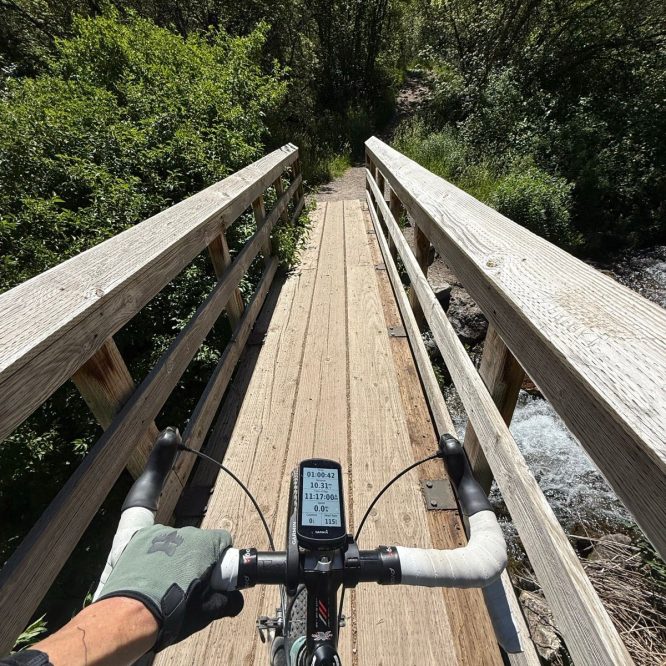 A cyclist's view from handlebars on a wooden bridge with trees and greenery on both sides, located in Providence, Utah.
