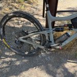 A close-up view of a mountain bike resting on a gravel path in Providence Canyon, Utah. The image shows the rear wheel and part of the bike's frame, highlighting the tire's treads and a water bottle attached to the bike. Surrounding the bike are patches of dry grass and small rocks.