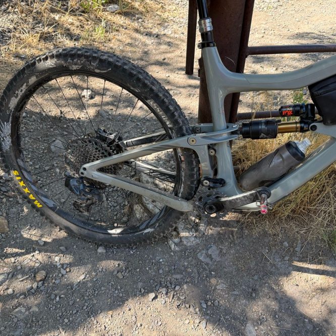 A close-up view of a mountain bike resting on a gravel path in Providence Canyon, Utah. The image shows the rear wheel and part of the bike's frame, highlighting the tire's treads and a water bottle attached to the bike. Surrounding the bike are patches of dry grass and small rocks.