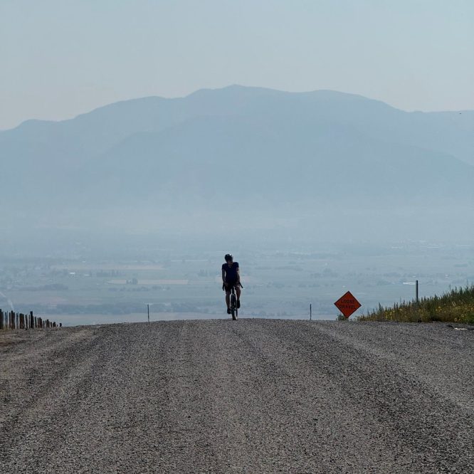 A person rides a bicycle on a gravel road leading into a hazy valley, with mountains in the background. A warning sign for loose gravel is visible on the right side. The scene is set in Logan, Utah, United States.