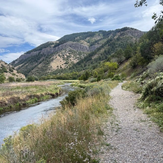 A gravel path winds through tall grass and shrubs alongside a stream, with mountainous terrain in the background under a partly cloudy sky. The scene is set in Providence, Utah, showcasing the area's natural beauty.